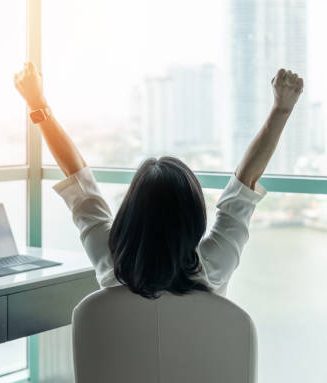 Business achievement concept with happy businesswoman relaxing in office or hotel room, resting and raising fists with ambition looking forward to city building urban scene through glass window